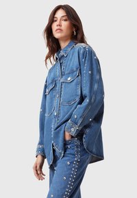 Camicia di denim blu con borchie argentate lungo le spalle, le tasche e le maniche. Presenta due tasche anteriori e un design con bottoni.