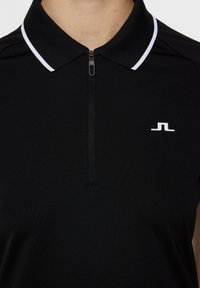 Svart polo-tröja med korta ärmar, med en dragkedja i halsen och en vit accent på kragen. Inkluderar en liten vit logotyp på bröstet.