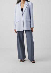 Světle modrý blazer s jednopákovým zapínáním, klopovými límci a dvěma předními kapsami, kombinovaný se šedými širokými kalhotami a černými sandály na blokovém podpatku.