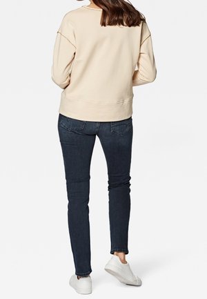 Femme portant un haut beige à manches longues, un jean skinny bleu foncé et des baskets blanches, debout dos au spectateur.