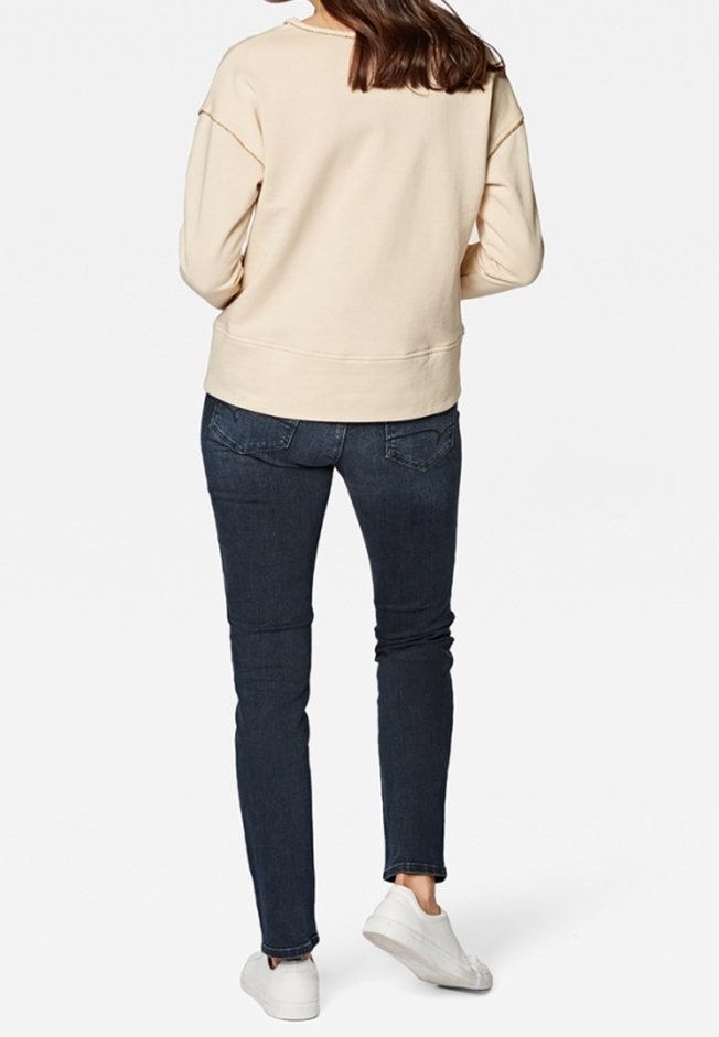 Femme portant un haut beige à manches longues, un jean skinny bleu foncé et des baskets blanches, debout dos au spectateur.