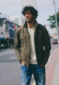 Olivgrüne Jacke mit Stehkragen, Knopfverschluss und zwei Brusttaschen, getragen über einem hellen Hemd und blauer Jeans.