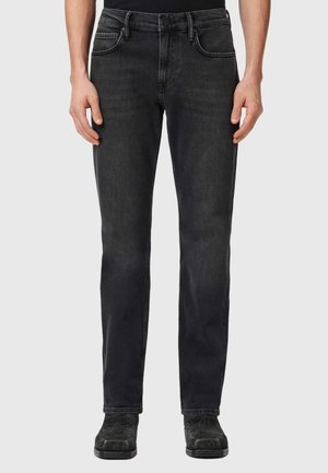 Man draagt zwarte slim-fit jeans en zwarte schoenen, staand tegen een effen lichtgrijze achtergrond.