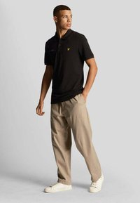 Polo nero con due bottoni, con marchio discreto sul petto, abbinato a pantaloni beige a gamba larga e sneakers bianche. Texture liscia.