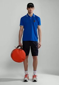 Mladý muž v modrom polo tričku, čiernych šortkách, čiernej čiapke a bielych teniskách s oranžovými šnúrkami, ktorý drží okrúhlu oranžovú tašku pri boku.