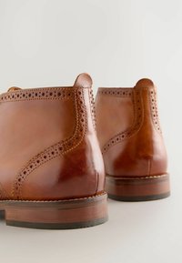 Brązowe skórzane botki z detalami w stylu brogue, okrągłymi czubkami i na wysokim obcasie. Gładkie wykończenie z kontrastującymi teksturami z perforacji i przeszyć.