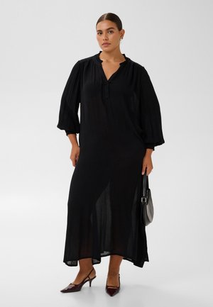 Modèle portant une longue robe noire à manches bouffantes, un petit sac à main noir et des escarpins bordeaux.