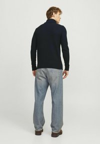 Pull en maille bleu marine avec un col haut, des poignets et un ourlet côtelés. Assorti à un jean bleu clair à coupe ample et des chaussures marron. Fond uni.