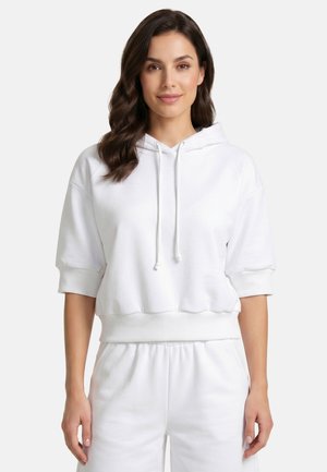 Frau mit welligem braunem Haar, die einen weißen kurzärmligen Hoodie und passende weiße Hose trägt, steht vor einem einfarbigen Hintergrund.