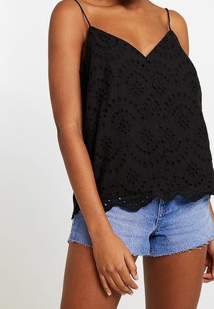 Czarna, bezrękawnikowa bluzka z ażurowym wzorem koronki i falowanym dołem, noszona z jasnoniebieskimi szortami dżinsowymi na stojącej osobie.