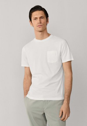 Hombre de pie con las manos en los bolsillos, vistiendo una camiseta blanca lisa de manga corta con un bolsillo en el pecho y pantalones gris claro.