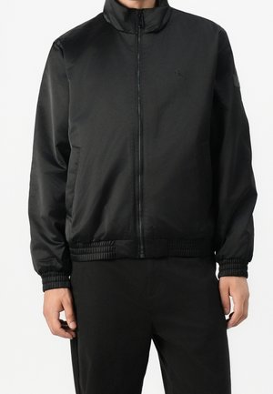 Homme portant une veste noire zippée avec des poignets et un ourlet élastiques, debout devant un fond blanc.