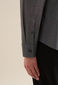 Närbild av en persons vänstra hand som vilar vid sidan, iklädd en långärmad grå skjorta med knäppta manschetter och svarta byxor.