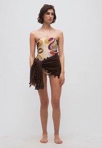 Γυναίκα που στέκεται ξυπόλυτη, φορώντας κορμάκι χωρίς τιράντες με σχέδια και σκούρα καφέ φούστα τύλιγμα δεμένη στη μέση, μπροστά σε απλό φόντο.