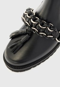 Μαύρο δερμάτινο παπούτσι με χοντρή σόλα, διακοσμημένο με πλεγμένο δέρμα και ασημένια αλυσίδα και δύο δερμάτινες φούντες στην μπροστινή πλευρά.