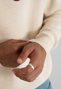 Mâini cu un ton de piele mediu-închis ajustând manșeta unei pulover din tricot crem, purtând un inel argintiu pe degetul mijlociu stâng.
