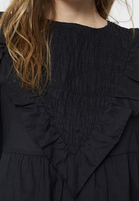 Robe noire avec une texture froncée au niveau de l'encolure, des manches évasées, et un détail en volants triangulaires sur le devant.