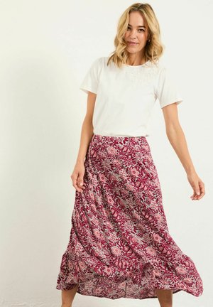 Frau mit blonden Haaren, die ein weißes Kurzarm-Oberteil und einen fließenden roten Blumenrock trägt, stehend vor einem schlichten hellen Hintergrund.