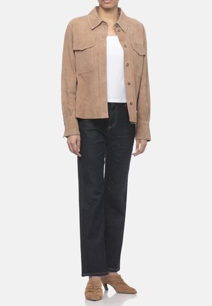 Persona che indossa una giacca beige con bottoni sopra una maglietta bianca, jeans neri a gamba dritta e scarpe beige con tacco arricciato, in piedi davanti a uno sfondo bianco.