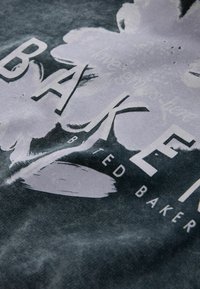 Tumši pelēks audums ar ziedu attēlu un vārdu „BAKER” treknā baltā fontā, ar teksturētu, nolietotu izskatu.