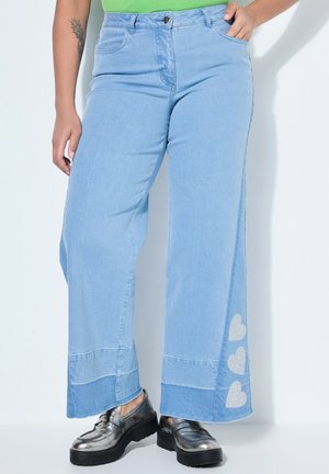 Hellblaue Jeans mit weitem Bein und drei silbernen Herz-Patches am unteren Bein, kombiniert mit metallisch-silbernen Plateauloafers und einem grünen Oberteil.