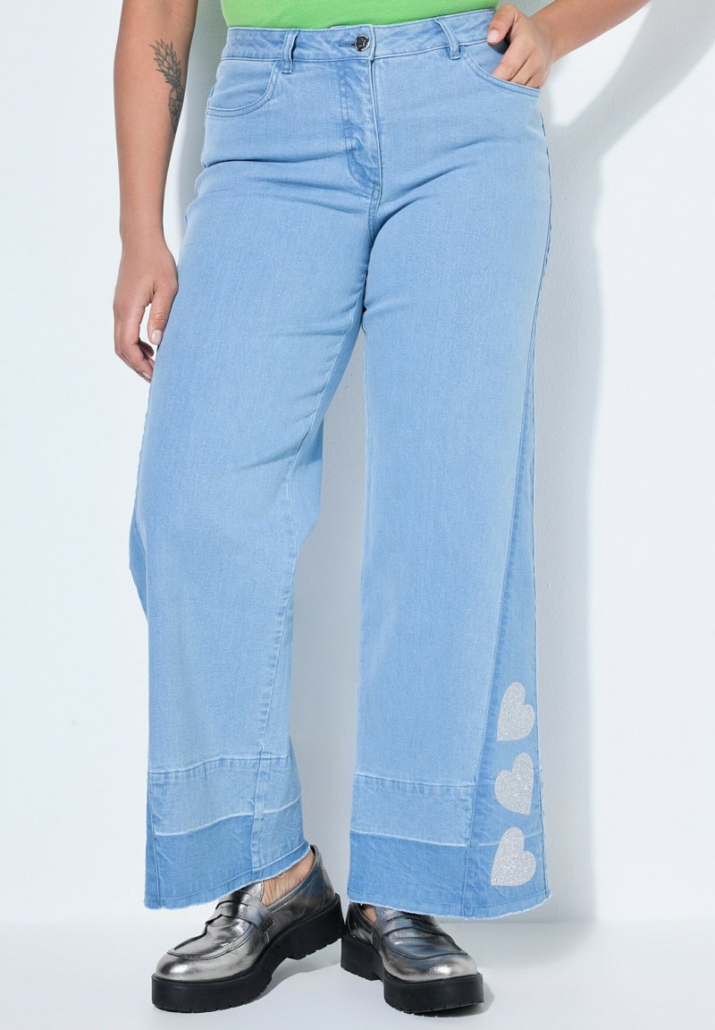 Jean large bleu clair avec trois patchs en forme de cœur argentés sur le bas de la jambe, porté avec des mocassins à plateforme argent métallisé et un haut vert.