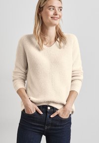 Cremefarbener Strickpullover mit V-Ausschnitt und lockerer Passform, ausgestattet mit gerippten Bündchen und einem strukturierten Muster, kombiniert mit dunklen Jeans.