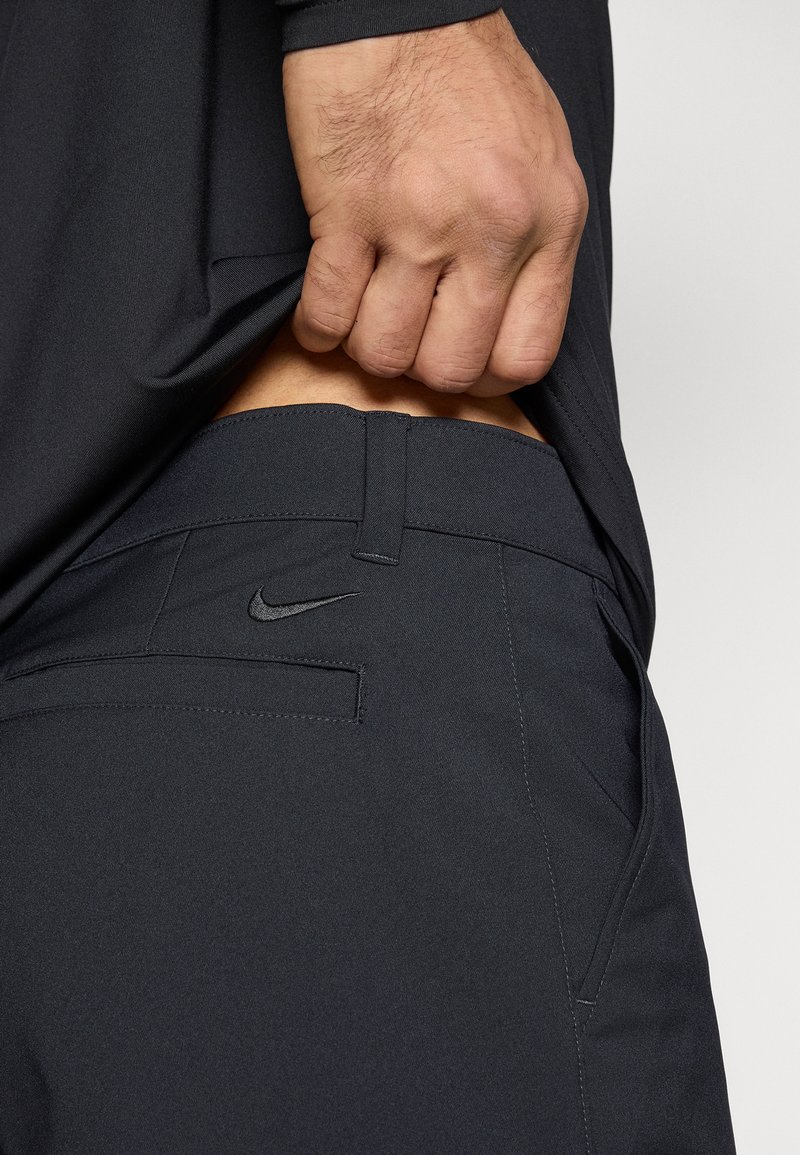 Schwarze Nike-Hosen mit einem eleganten Design, die ein dezentes Logo, strukturierten Stoff und versteckte Taschen aufweisen, sind vor einem grauen Hintergrund präsentiert.
