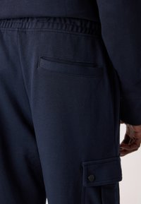 Gros plan d’un pantalon bleu marine avec taille élastique, poche passepoilée à l’arrière et poche cargo inférieure avec rabat boutonné sur le côté droit d’une personne.