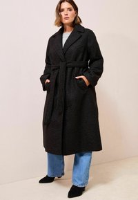 Sort uldblandet frakke med bælte, tekstureret stof, spidse slag og sidelommer, parret med lyseblå jeans og sorte spidse sko.