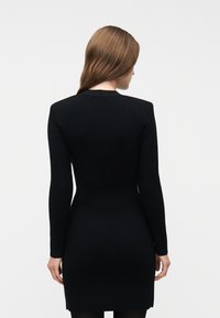 Γυναίκα με μακριά καστανά μαλλιά φοράει εφαρμοστό μαύρο πλεκτό φόρεμα με μακριά μανίκια, στραμμένη πλάτη σε απλό φωτεινό φόντο.