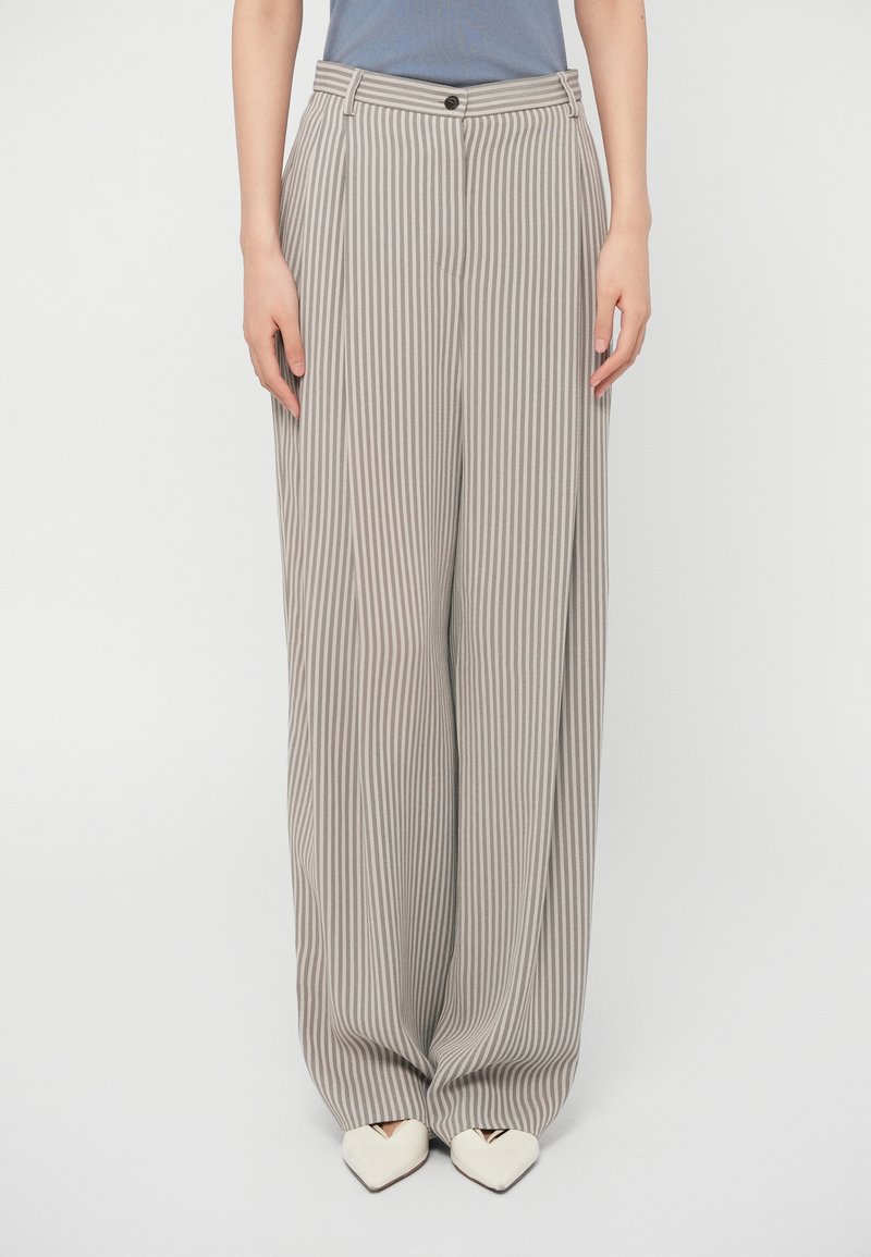 Beżowo-szare szerokie spodnie w paski, z wysokim stanem, fałdami z przodu i kieszeniami po bokach. Stylizowane z białymi, spiczastymi butami.