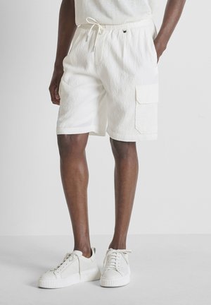 Uomo che indossa pantaloncini bianchi strutturati con tasca laterale e scarpe da ginnastica bianche con lacci, in piedi su un pavimento bianco semplice.