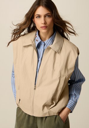 Donna con capelli castani ondulati indossa un gilet beige senza maniche con cerniera sopra una camicia a righe blu e marroni con bottoni, con le mani nelle tasche dei pantaloni verde oliva.