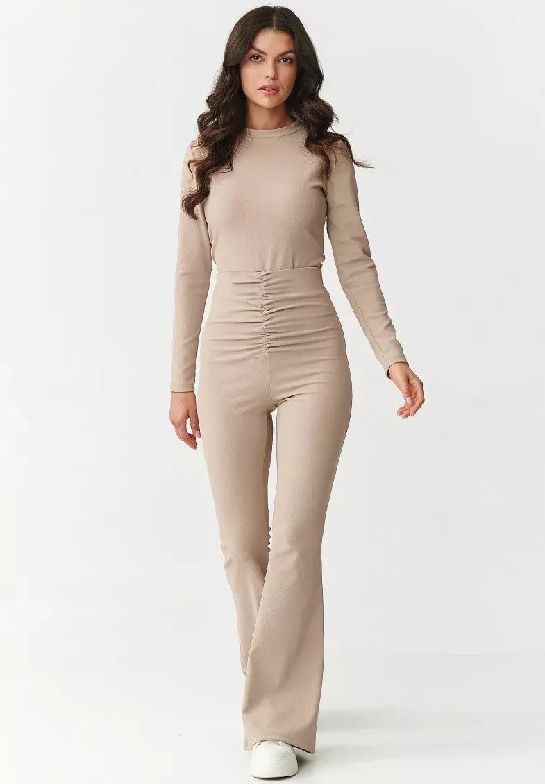 Beżowy kombinezon z długim rękawem, z dopasowanym gorsetem i marszczeniem w talii; miękka prążkowana faktura przechodząca w rozszerzane nogawki; w połączeniu z białymi butami.