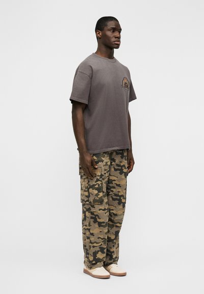 Γκρι βαμβακερό μπλουζάκι με κοντά μανίκια και λογότυπο μπροστά, συνδυασμένο με φαρδιά παντελόνια καμουφλάζ τύπου cargo. Το μοντέλο φοράει ανοιχτόχρωμα αθλητικά παπούτσια.