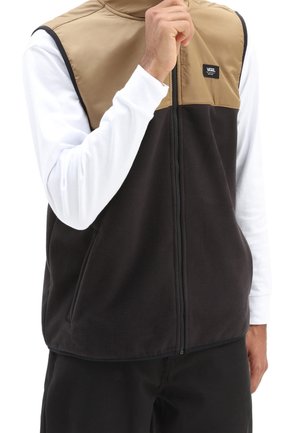 Sort fleece vest med en tan nylon overdel, frontlynlås og to sidelommer. Bæres over en langærmet hvid skjorte med et logo-patch.