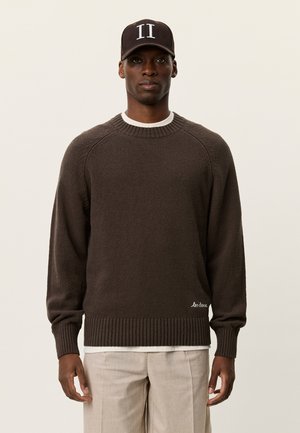 Donkerbruine gebreide trui met geribde manchetten en zoom. Heeft een ronde halslijn en een geborduurd logodetail onderaan.