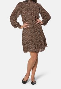 Braunes gemustertes Kleid mit hochgerüschtem Halsauschnitt, langen Puffärmeln und einem Rüschensaum; getragen mit schwarzen spitzen Pumps.