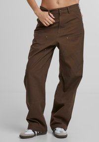 Braune weit geschnittene Hose aus Baumwolle mit metallischen Akzenten, großen Vorderentaschen und lockerer Passform, kombiniert mit weißen und grauen Sneakers.