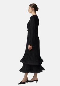 Robe plissée noire à manches longues et ourlet en tiers fluide, fabriquée en tissu léger. Associée à des chaussures noires à bout pointu.