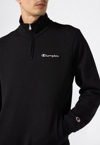 Uomo che indossa un felpa nera Champion con zip corta, con logo ricamato bianco sul petto e piccolo logo sul polsino sinistro.