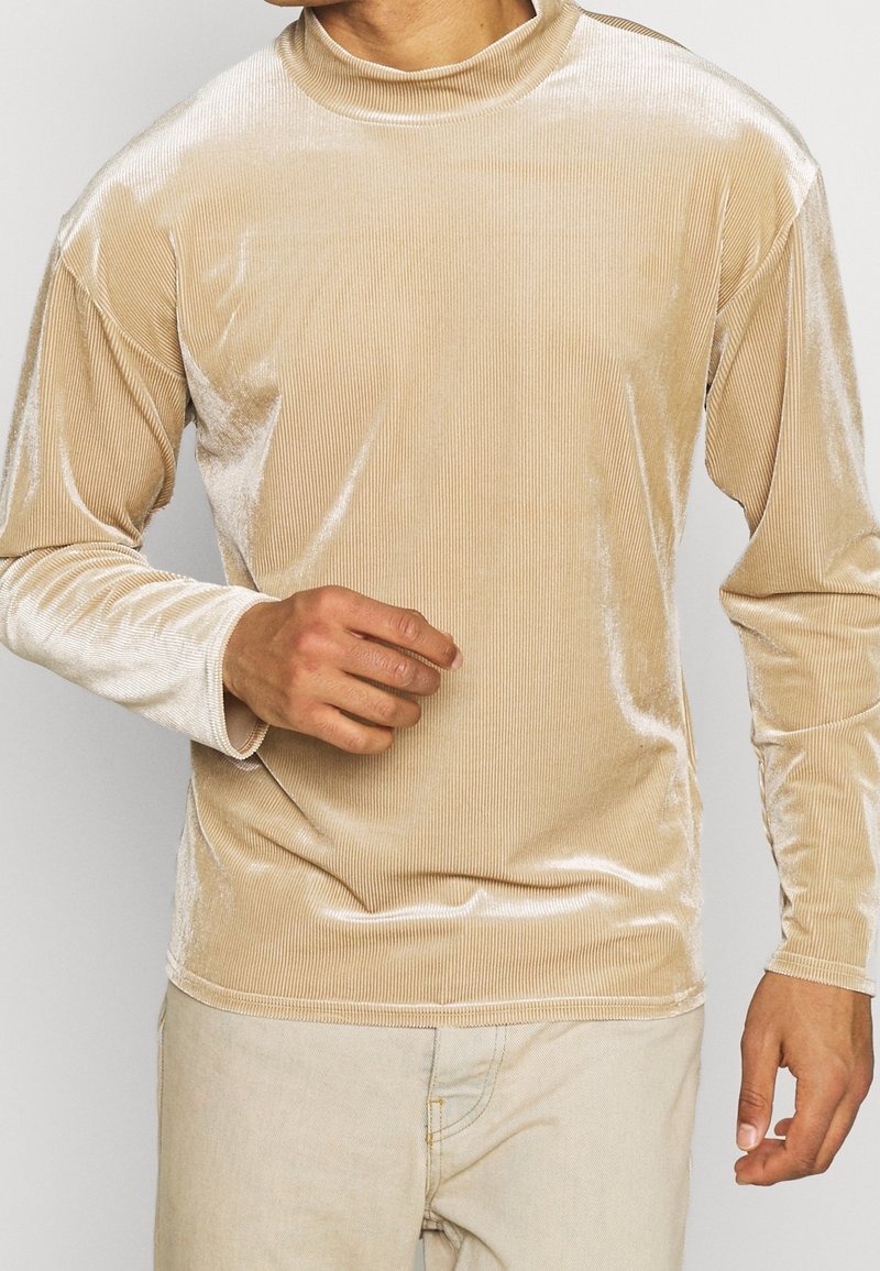 Homme portant une chemise à manches longues en velours beige et un pantalon beige clair, vu du cou jusqu'aux hanches avec une main légèrement levée.