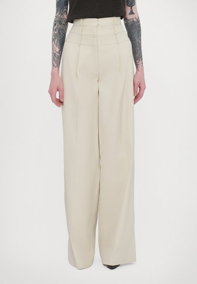 Pantalons beiges à taille haute, jambes larges avec plis, fabriqués en tissu lisse, présentant des coutures ajustées et une silhouette propre et structurée.