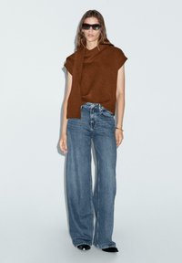 Top in maglia corto maniche, di colore marrone, con dettagli attorcigliati, abbinato a jeans a gamba larga di colore blu chiaro. Accessoriato con occhiali da sole neri e un bracciale d'oro.