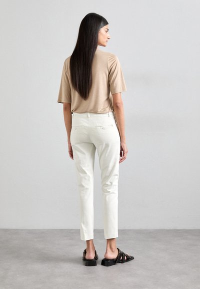 Cămașă bej cu mânecă scurtă, pantaloni albi ajustați și sandale negre cu toc pătrat. Textură netedă, design minimalist, cu buzunare pe spate vizibile.