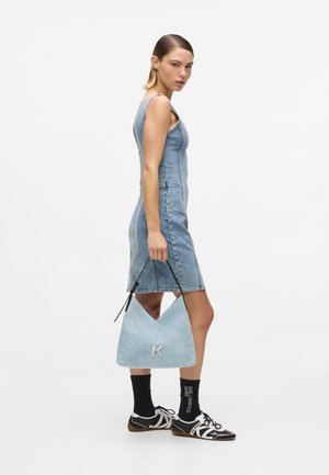 Kvinne i ermeløs denimkjole holder lyseblå håndveske med «K»-logo, iført svarte sokker med hvit skrift og svart-hvite joggesko.