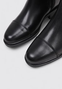 Botines de cuero negros con una textura suave, puntera redondeada, costuras prominentes y un tacón bajo. Diseño sencillo sin acentos.