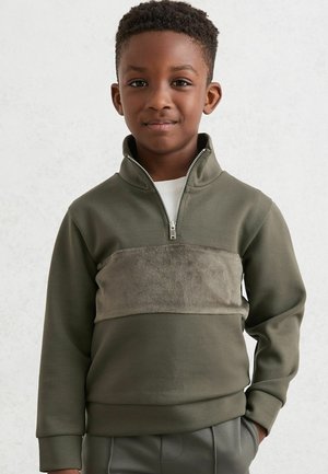 Olive zaļas krāsas zip-up sporta krekls ar mīkstu, bēšas krāsas flīsa paneli uz krūtīm. Garas rokas, augsta apkakle un ribota aproce.