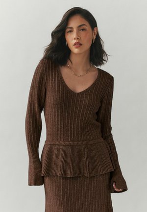 Nainen, jolla on aaltoilevat tummat hiukset, pukeutuneena ruskeaan ribbineulottuun peplumtunikaan ja vastaavaan hameeseen, kullanvärisiin korvakoruihin ja ohueen kultakaulakoruun.